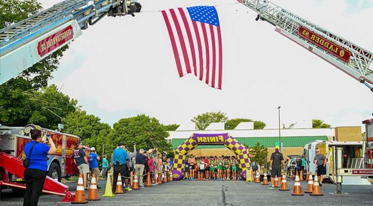 A photo of the Run White and Blue event's finish line in the distance with a cluster of racers behind it. An American flag is being flown by two firetruck ladders in the foreground and the angle of the photo shows the flag being directly above the finish line.